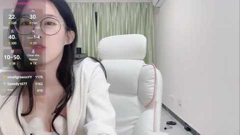 Media: Video of a young Asian woman with fair skin, long black hair, and glasses, wearing a white cardigan, sitting on a white leather chair in a modern, minimally decorated room.