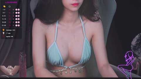 Media: A video of an Asian woman with long dark hair, wearing a light blue bikini top, sitting on a gray chair. Background includes a dimly lit room with a purple neon drawing on the wall.