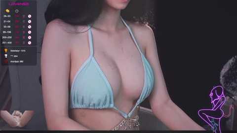 Video of a young woman with long black hair, wearing a low-cut, light blue halter bikini top, exposing ample cleavage, and a metallic necklace. The background shows a dark, dimly lit room with a purple neon outline of a woman's figure.