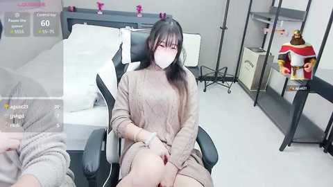 Media: Video of an Asian woman in a beige sweater, sitting on a black chair in a sterile room, wearing a face mask.