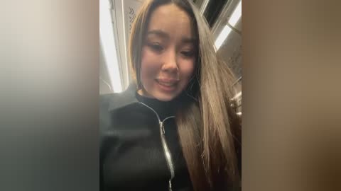 Media: Video of a young woman with light skin and long, straight blonde hair, smiling and looking down, wearing a black jacket. She is in a dimly lit subway car, with fluorescent lights and graffiti on the walls.