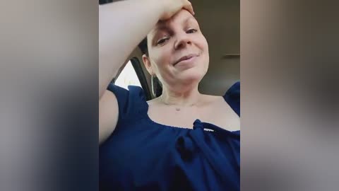 Media: Video of a smiling, fair-skinned woman with dark hair, wearing a dark blue top, sitting in a car, holding her head with one hand.