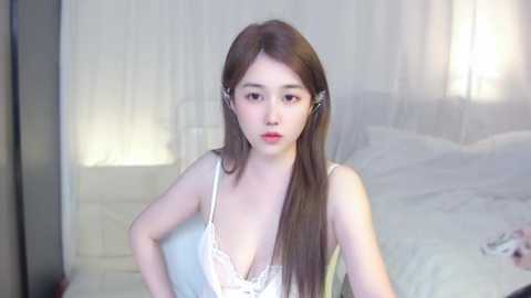 Media: A video of a young Asian woman with long brown hair, fair skin, wearing a white lace lingerie top, standing in a dimly lit bedroom with white curtains and a bed.