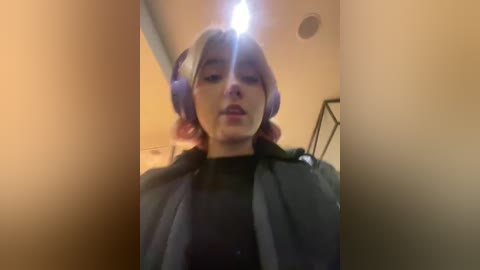 Media: Video of a young woman with pink hair wearing large headphones, black turtleneck, and a gray coat, standing in a dimly lit hallway with beige walls.