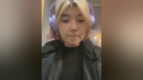 Media: A video of a young Asian woman with light skin, platinum blonde hair, and large purple headphones, wearing a black jacket, looking down in a dimly lit room.