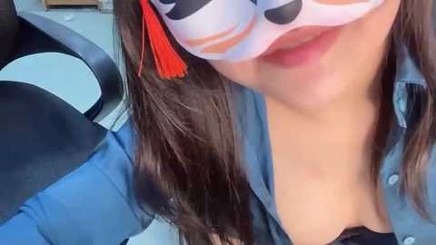 Media: Video of a woman with light skin, long dark hair, and a white mask with orange accents, wearing a blue shirt. She's seated in a chair with black upholstery.