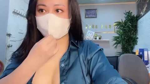 Media: Video of a young woman with long black hair, wearing a blue shirt and a white face mask, holding her chin, in a modern office with a plant, clock, and framed art in the background.