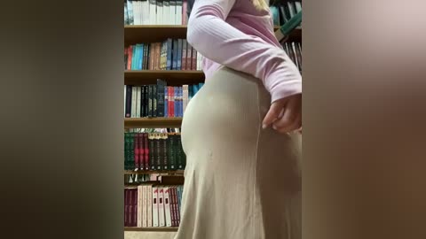 Media: Video of a person with light skin, wearing a pink long-sleeve top and beige dress, standing in a room with wooden bookshelves filled with books.