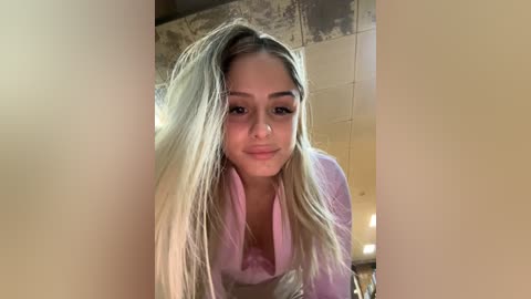 Media: Video of a young woman with long, straight blonde hair, wearing a light pink shirt, leaning forward. She has a light complexion and is smiling. The background features beige walls with a slightly worn texture.