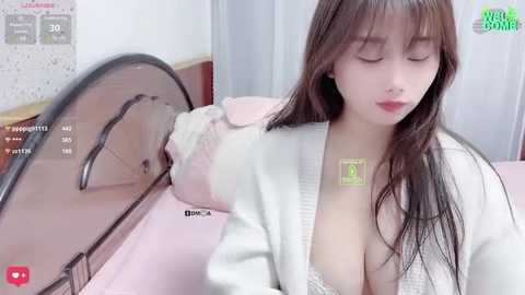 Media: Video of an East Asian woman with long brown hair and fair skin, lying on a pink bed with a white cardigan open, showing cleavage, in a modern bedroom with a white wall.