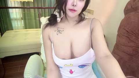 Media: Video of an East Asian woman with long black hair, wearing a revealing white tank top with large cleavage, sitting in a bedroom with a bed and green curtains.