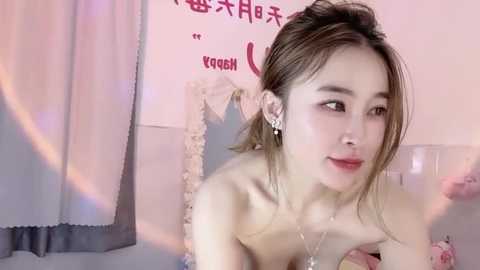 Media: Video of a young Asian woman with fair skin, brown hair tied back, wearing a strapless top and dangling earrings, looking contemplative. Background features pink and white decor with Chinese text.