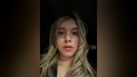 Media: Video of a young, light-skinned woman with long, wavy, blonde hair, wearing minimal makeup and a black top, set against a dark, blurry background.