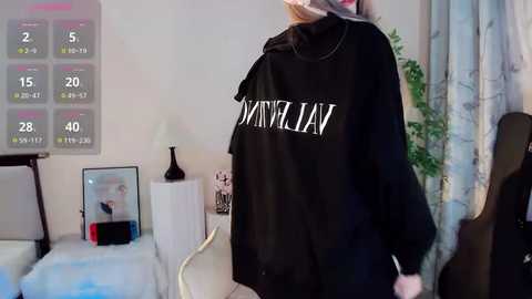 Media: Video of a woman in a black oversized turtleneck sweater with \"OWN YOUR OWN\" in white, standing in a cozy bedroom with a white bed, potted plant, and calendar on the wall.