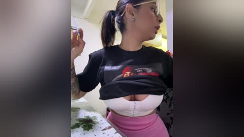 Media: Video of a Latina woman with light skin and long dark hair in a ponytail, wearing a black t-shirt with a graphic design, and a white bra exposing her cleavage, pink shorts, and a tattooed arm, posing indoors.