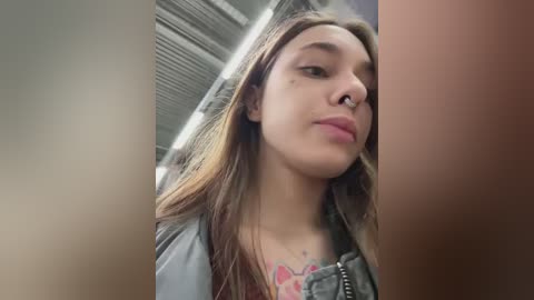 Media: A video of a young woman with light skin and long, wavy brown hair, wearing a gray leather jacket and a floral top, captured in an indoor setting with metal ceiling beams.