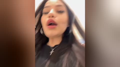 Media: A video of a woman with long, dark hair, light skin, and full lips, wearing a black top, standing indoors with a blurred background.