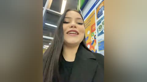 Media: Video of a smiling young woman with long black hair, wearing a black blazer, standing in a brightly lit convenience store. Background features colorful posters and shelves.