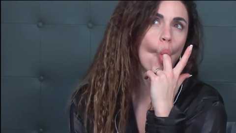 Media: Video of a woman with long, curly brown hair, light skin, and blue eyes, holding her finger to her lips, wearing a black jacket, against a gray tufted headboard background.