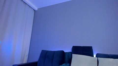 Media: Video of a minimalist living room corner featuring a grey couch with beige cushions, against a light grey wall, with a blue ambient light highlighting the room's modern, neutral decor.