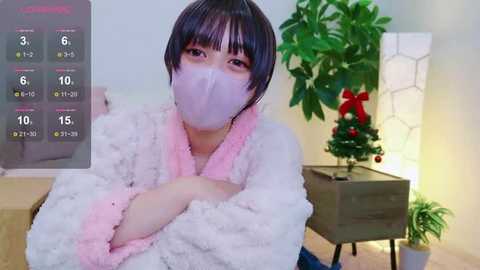 Media: Video of an East Asian woman with short black hair, wearing a pink mask, fluffy white robe, and blue jeans, seated in a room with a Christmas tree and potted plant, calendar overlay.