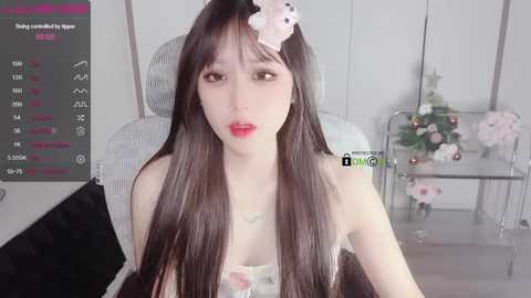 Media: Video of a young Asian woman with long black hair, pale skin, and red lipstick, wearing a white floral top, seated in a white chair with a glass table behind her.