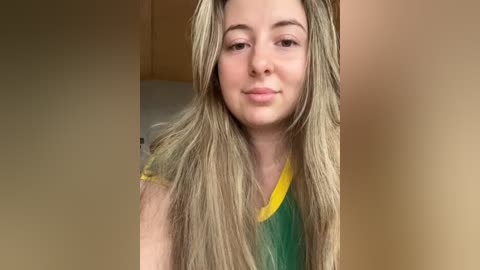 Media: Video of a young Caucasian woman with long, straight blonde hair, wearing a green and yellow T-shirt, standing in front of a beige wall, smiling softly.