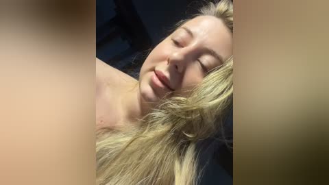 Media: Video of a fair-skinned, blonde woman with closed eyes, lying on a bed, her hair messy, against a dark, blurred background.