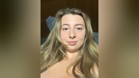 Media: Video of a young, light-skinned woman with long, wavy blonde hair, smiling, topless, in a dimly lit bedroom with wooden walls and a blue bedspread.