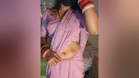 Media: Video of a woman in a purple sari, exposing her midriff, with red and white bangles, in a dimly lit room with string lights.