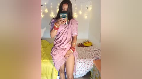 Media: A young South Asian woman with long dark hair, wearing a pink saree and red bangles, takes a selfie in a cozy bedroom with string lights and a bed with colorful bedding.