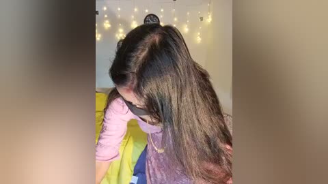 Media: Video of a woman with long black hair, wearing a purple shirt and black mask, leaning over a yellow couch, with warm fairy lights in the background.