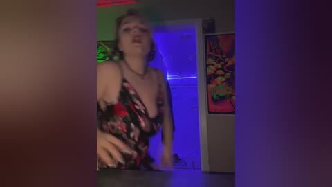 Media: Video of a curvy woman with shoulder-length blonde hair, wearing a black floral dress, in a dimly lit room with psychedelic posters and blue lighting.