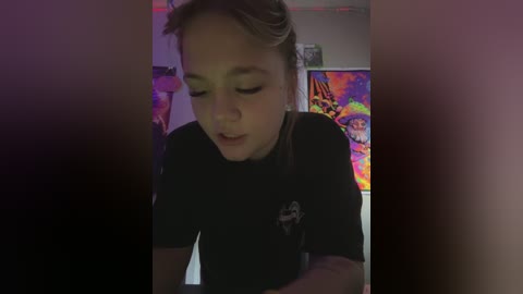 Media: A video of a young woman with light skin and blonde hair, wearing a black t-shirt, leaning forward, in a dimly lit room with colorful, abstract art on the wall.