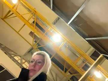 Media: A video of a blonde woman with glasses, wearing a black top, standing in an industrial setting with yellow scaffolding, metal beams, and bright lights.