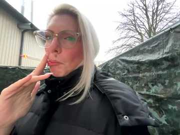 Media: Video of a blonde Caucasian woman with light skin, wearing clear glasses and a black jacket, standing outdoors near a chain-link fence and industrial building, with a bare tree in the background.