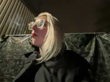 Media: Video of a blonde woman with large, transparent glasses, wearing a black coat, smoking a cigarette, standing in a dimly lit, industrial setting with a corrugated metal wall and plastic sheeting behind her.