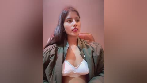 Media: A video of a young South Asian woman with long black hair, wearing a white bra and open green jacket, seated against a beige wall.