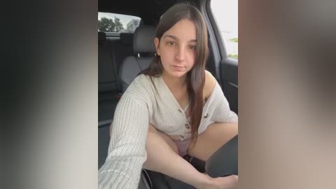 Media: Video of a young woman with long dark hair, fair skin, and brown eyes, wearing a light gray, off-shoulder knitted sweater, sitting in a car.