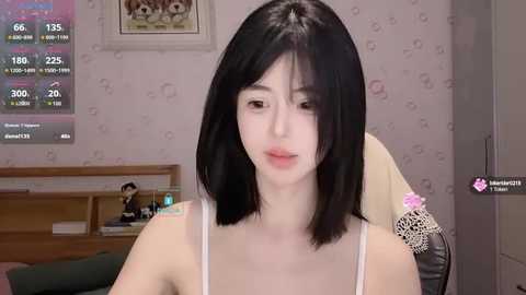 Media: Video of a pale-skinned Asian woman with shoulder-length black hair, wearing a white spaghetti-strap top, sitting indoors with a gaming headset. Background features a pink patterned wall, gaming monitor, and a framed picture.