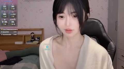 Media: Video of a young, East Asian woman with fair skin, dark hair, and a white robe, seated in a gaming chair in a bedroom with a bed, wooden shelves, and a TV screen displaying streaming statistics.