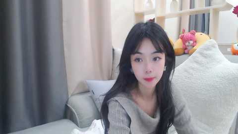 Media: Video of an Asian woman with long black hair, fair skin, and red lipstick, wearing a gray sweater, sitting on a beige sofa in a softly lit living room.