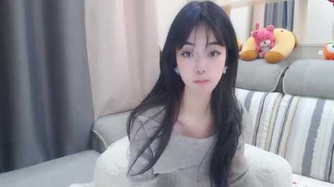 Media: Video of a young Asian woman with long black hair, fair skin, and delicate features, wearing a gray sweater, seated on a bed with striped pillows and stuffed toys in a cozy, softly lit room.