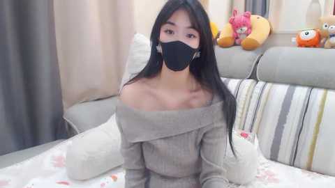 Media: A video of an East Asian woman with long black hair, wearing a black mask and off-shoulder gray sweater, seated on a bed with striped pillows and stuffed toys in the background.