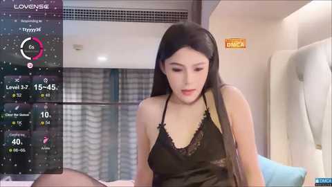 Media: Video of a young Asian woman with long black hair, wearing a black lace camisole, sitting on a bed in a modern bedroom.