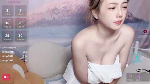 Media: A video of an East Asian woman with fair skin and medium-sized breasts, partially covered by a white towel, sitting on a bed with a pink and blue background.