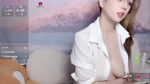 Media: Video of a young, fair-skinned woman with long brown hair, partially undressed in a white blouse, revealing her bare breasts. The background features a blurred, serene landscape.