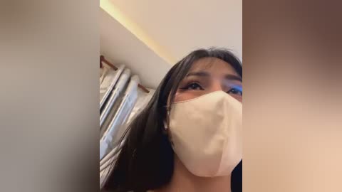 Media: A video shows an Asian woman with long black hair, wearing a beige surgical mask, standing in a room with beige walls and a window with light blue curtains.