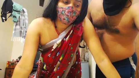 Media: Video of a shirtless man with a mustache, wearing a mask, leaning over a woman in a colorful saree. Background shows hanging clothes and a dark-haired woman.