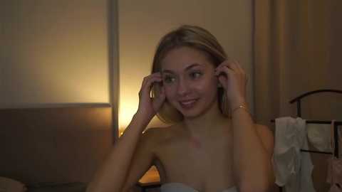 A video of a smiling, fair-skinned blonde woman with small breasts, adjusting her glasses in a dimly-lit, cozy bedroom with a nightstand and curtains in the background.